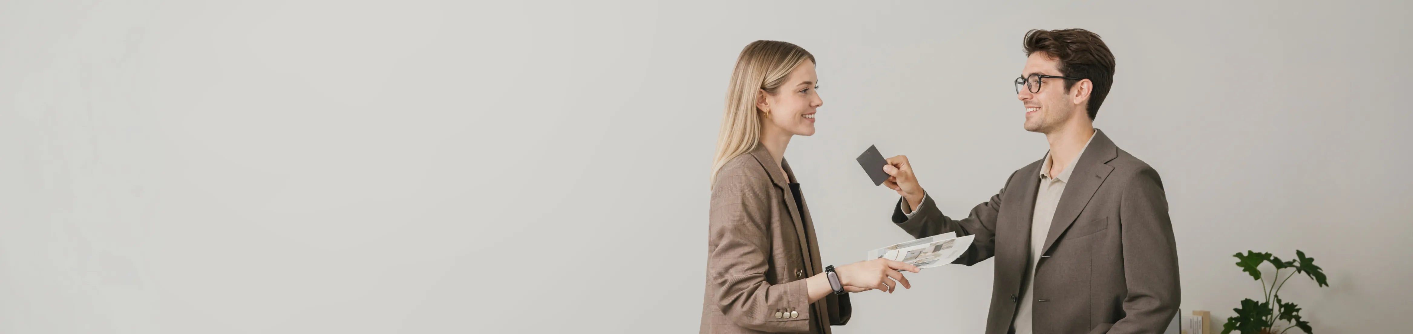 A smiling man in a brown blazer presents a dark Plaud Note Pro to a woman in a brown blazer holding papers.