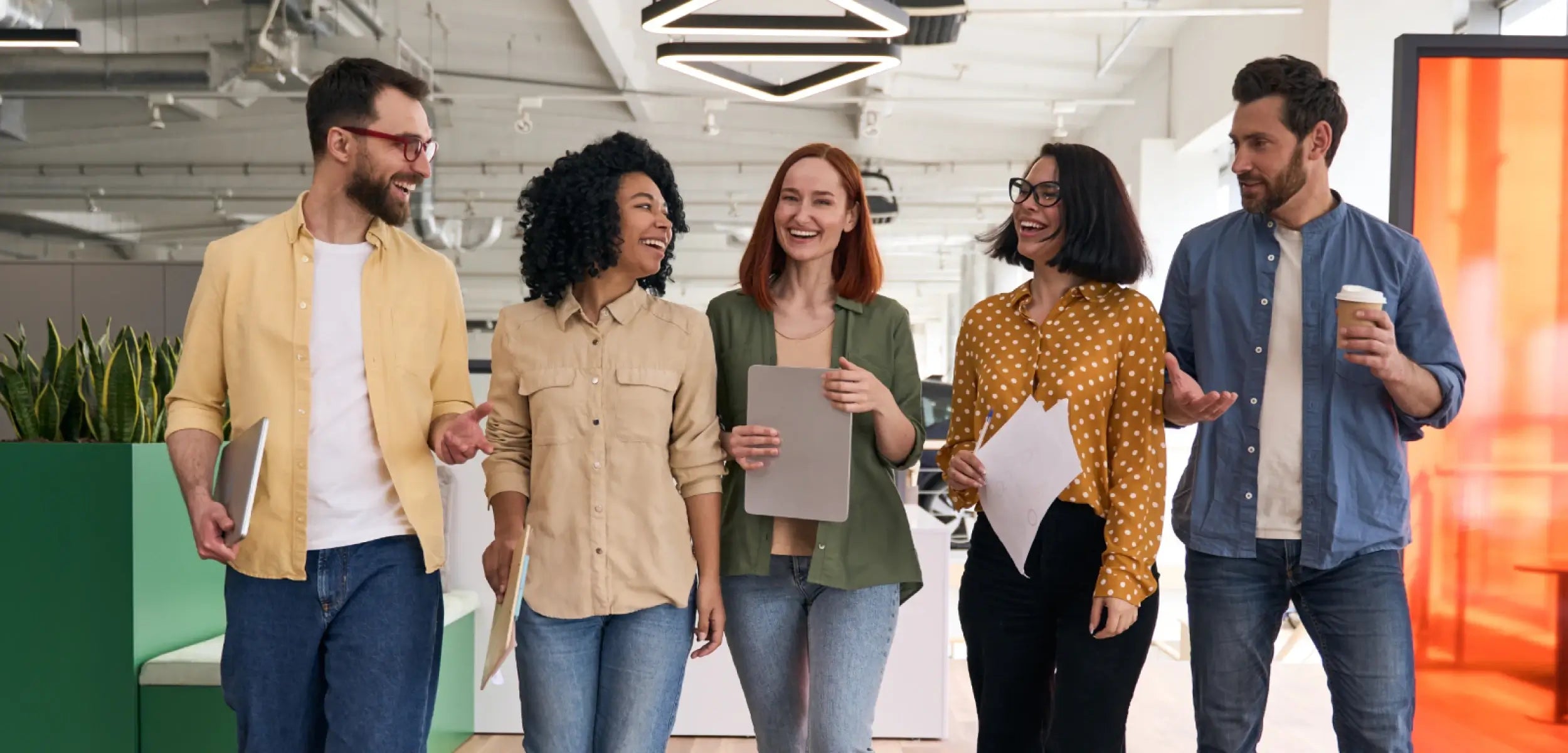 Five diverse colleagues walk and chat happily in a bright, modern office hallway, conveying a positive work environment.