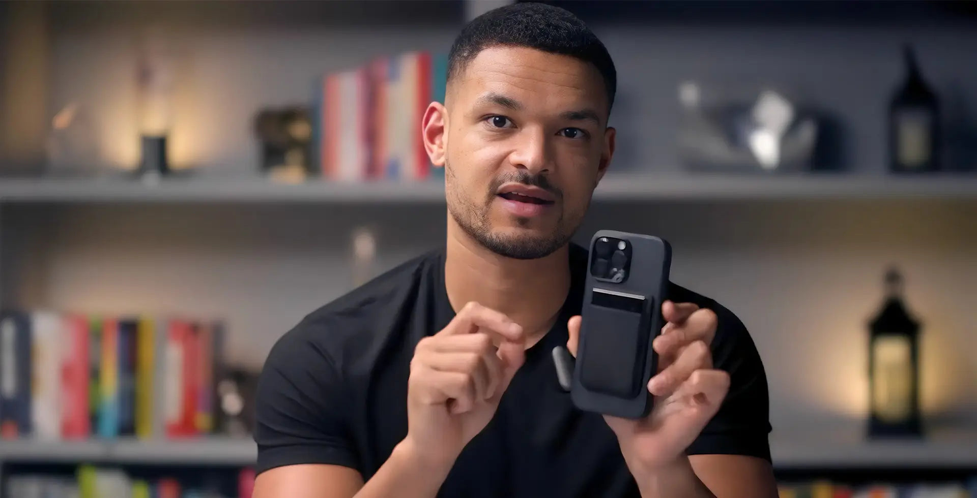 A man in a black shirt holds a black smartphone with a black Plaud Note Pro attached to its case, gesturing in front of a boo