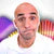 A bald man in a white shirt looks directly at the camera, with a blurred colorful background.