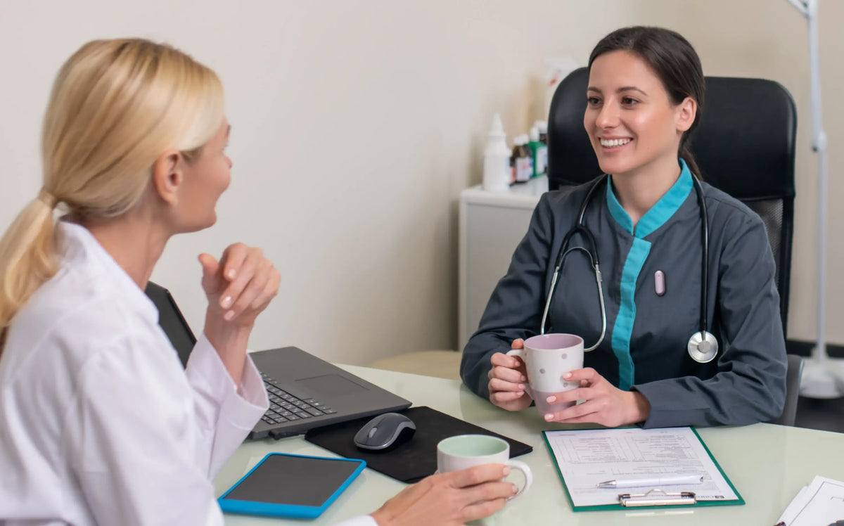 A smiling doctor in grey scrubs with a pink Plaud NotePin S clipped to her top, holds a mug while talking to a colleague in a