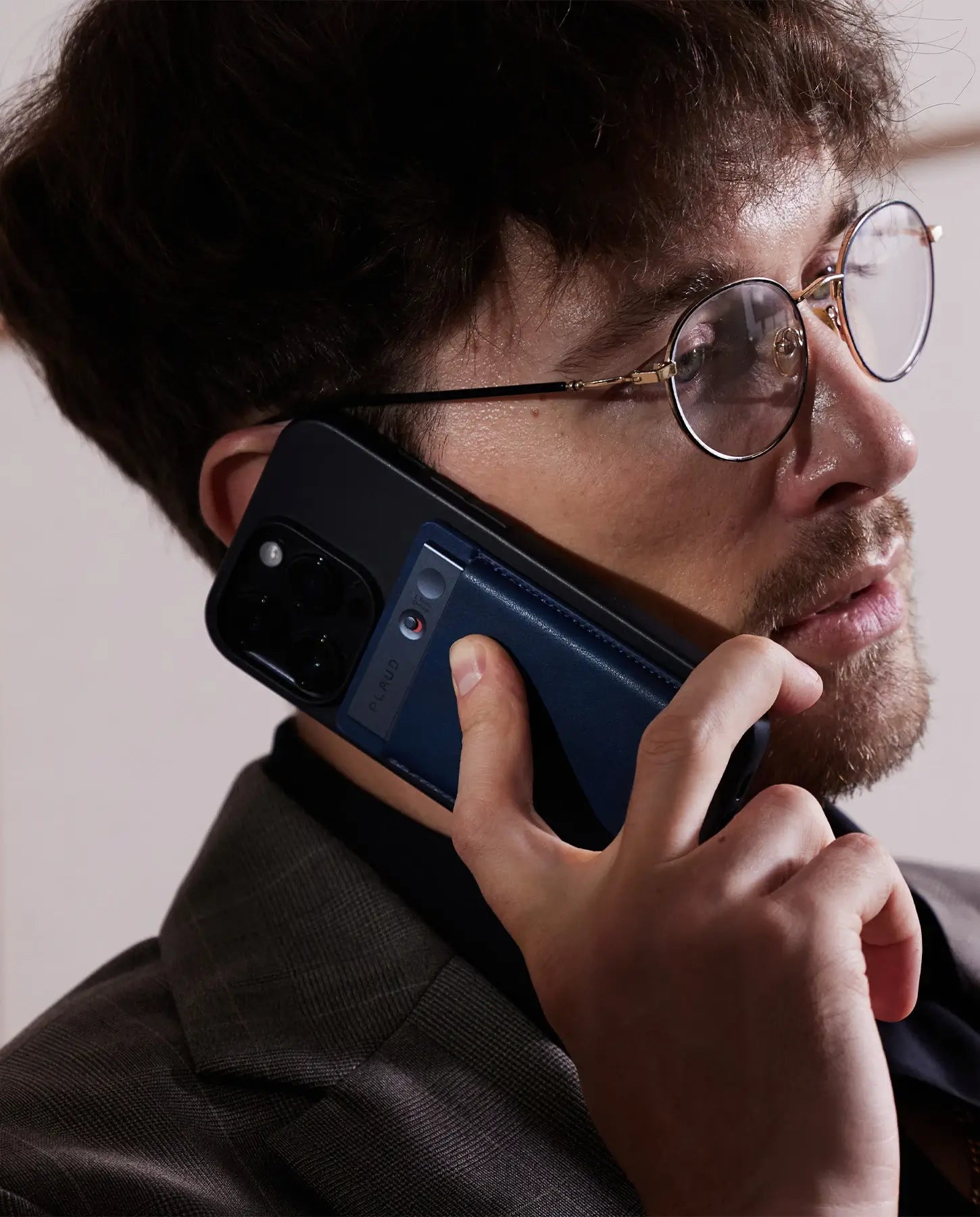 A man in glasses holds a phone to his ear, with a dark blue Plaud Note attached to the back.