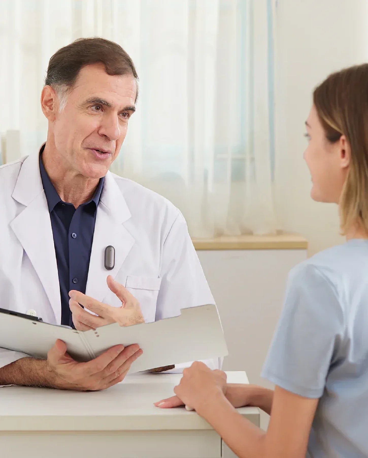 A male doctor in a white coat, with a dark grey Plaud NotePin clipped to his lapel, consults with a female patient.