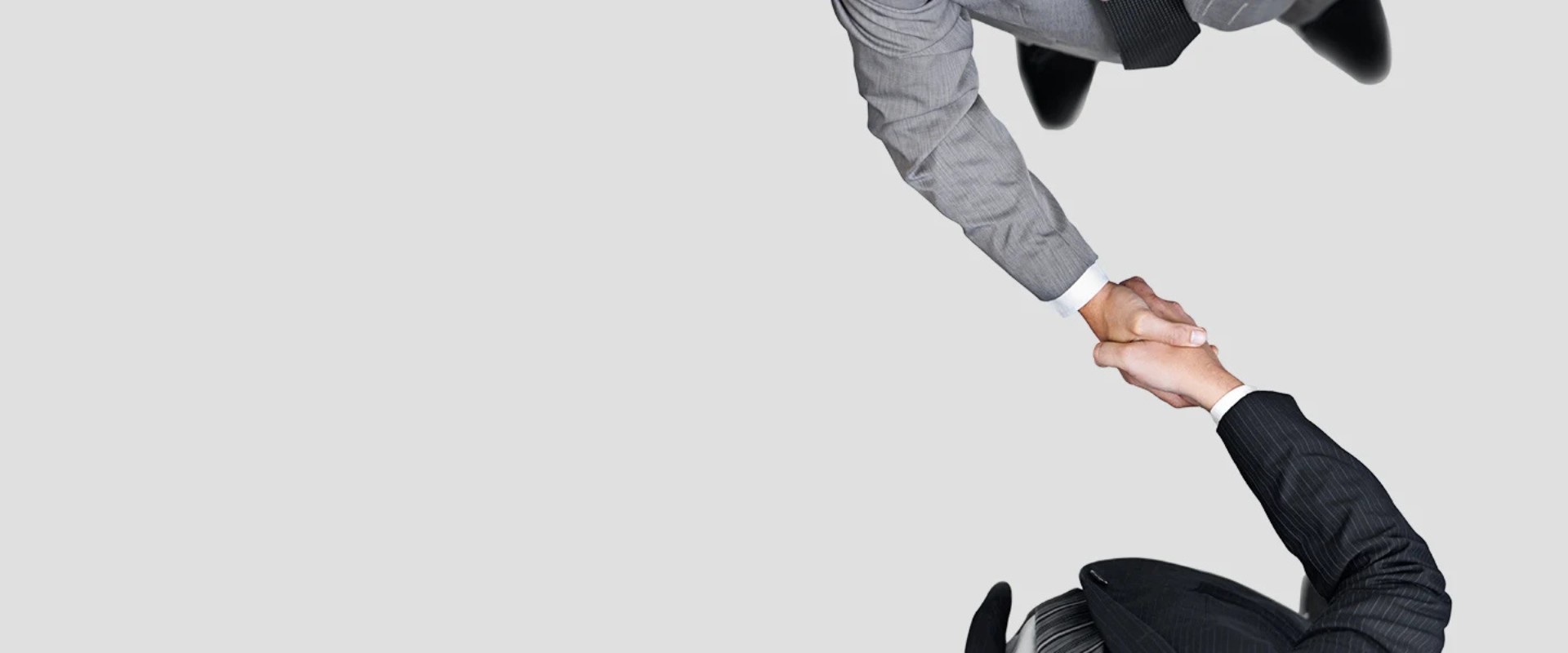 Two business people in suits shake hands, viewed from above, on a light grey background, conveying partnership.