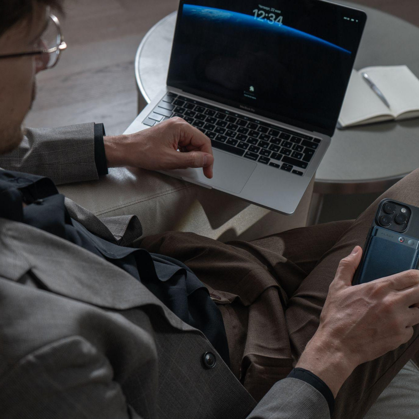 A man in a suit uses a laptop while holding a phone with a black Plaud Note AI voice recorder attached to the back.
