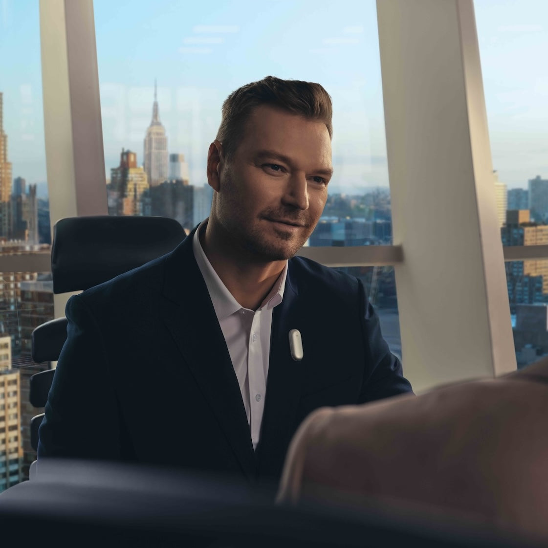 A man in a dark suit sits in a high-rise office with a city skyline view, a white Plaud NotePin clipped to his lapel.