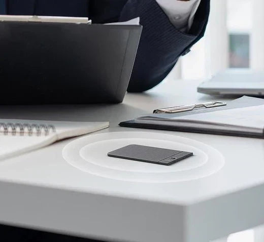 A dark gray Plaud Note lies on a white desk with white concentric circles suggesting active recording, in a professional sett