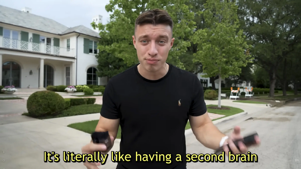 A man in a black shirt holds two black Plaud Note Pro devices, standing on a paved road in front of a large white mansion.