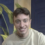 A young man with short brown hair and a light-colored sweatshirt smiles at the camera, with plants and a blue wall in the bac