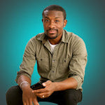 A man with dark skin and a beard, wearing a green shirt, sits holding a black smartphone against a teal background.