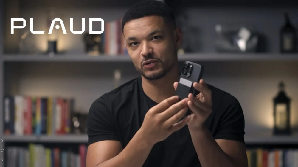 A man in a black t-shirt holds up a dark smartphone with a silver Plaud Note Pro magnetically attached to its back, in a blur