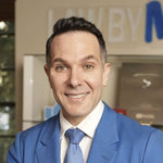 A smiling man with dark hair and light eyes, wearing a blue suit and tie, against a blurred office background.