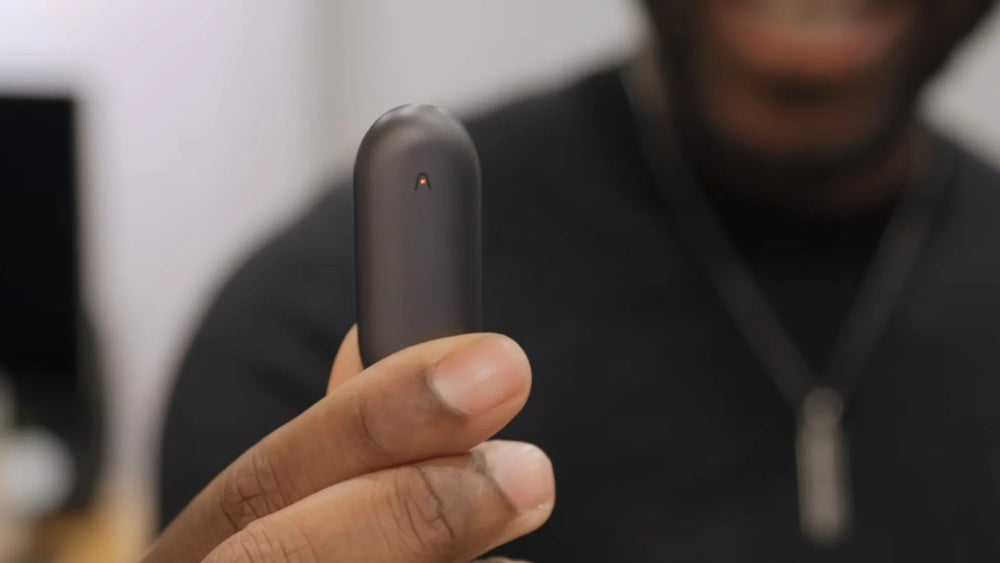 A person's hand holds A black Plaud NotePin, which has a small red indicator light on its face.