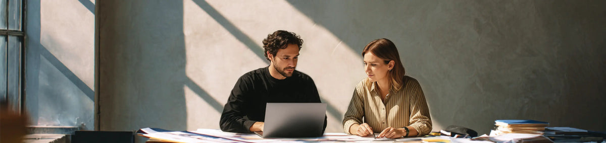 Two colleagues, a man and a woman, collaborate intently on a laptop at a sunlit desk in a modern office space.