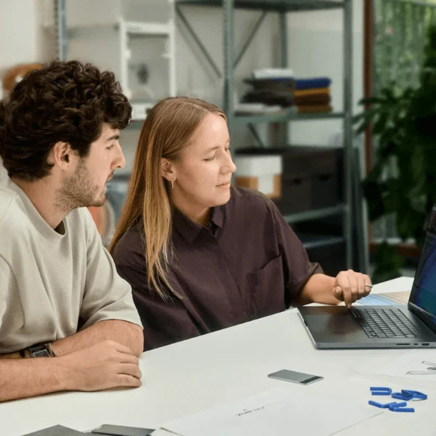 Two colleagues collaborate at a white desk, with a silver Plaud Note resting nearby, as they review content on a laptop.