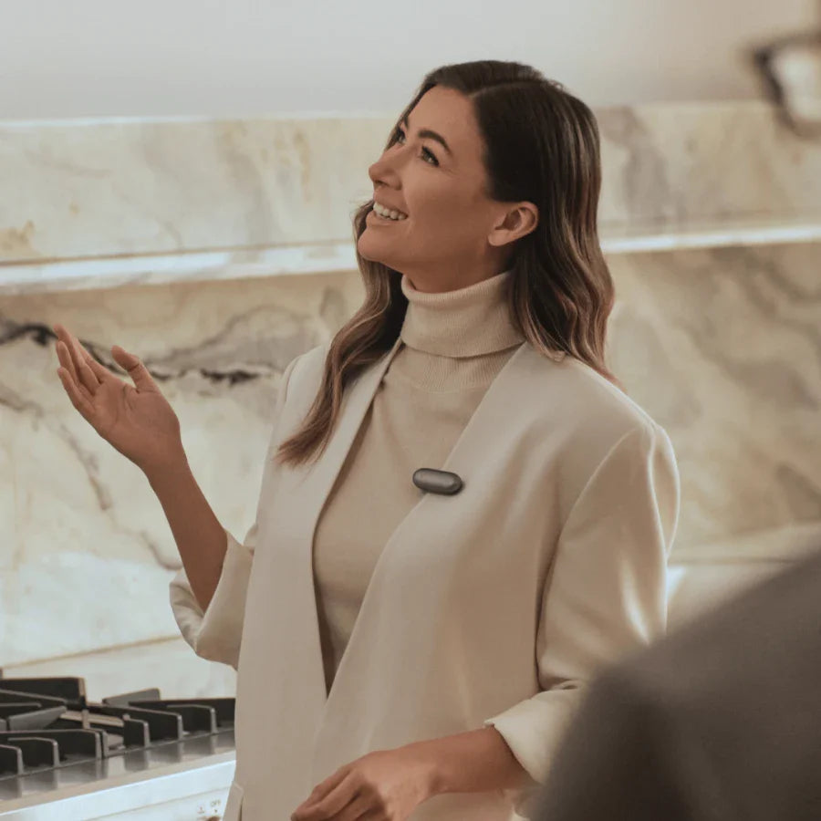 A smiling woman in a cream blazer and turtleneck wears a dark gray Plaud NotePin clipped to her lapel in a modern kitchen set