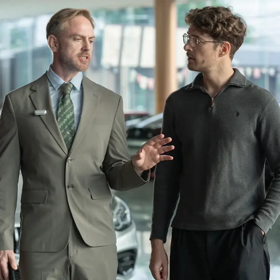 A salesman in a suit with a silver Plaud NotePin clipped to his lapel talks to a customer in a car dealership.