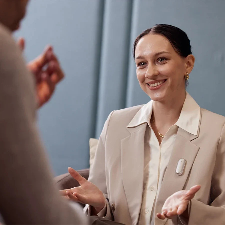 A smiling woman in a light blazer gestures while conversing, with a white Plaud NotePin S clipped to her lapel.