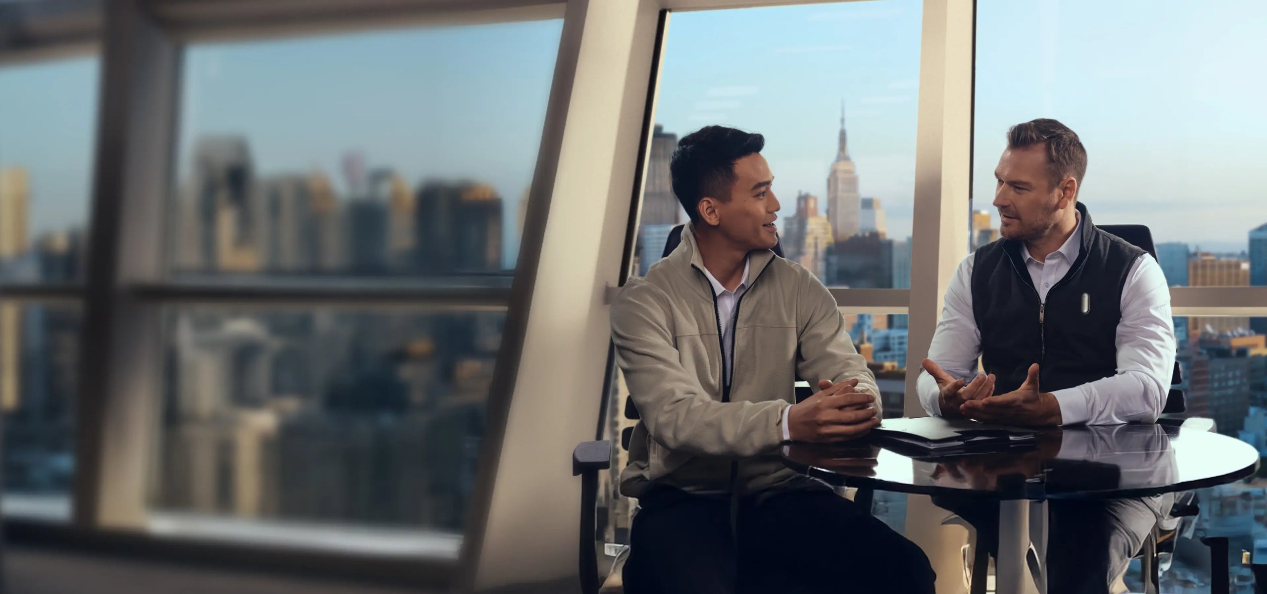 Two men converse at a round table in a high-rise office overlooking a city skyline with the Empire State Building.