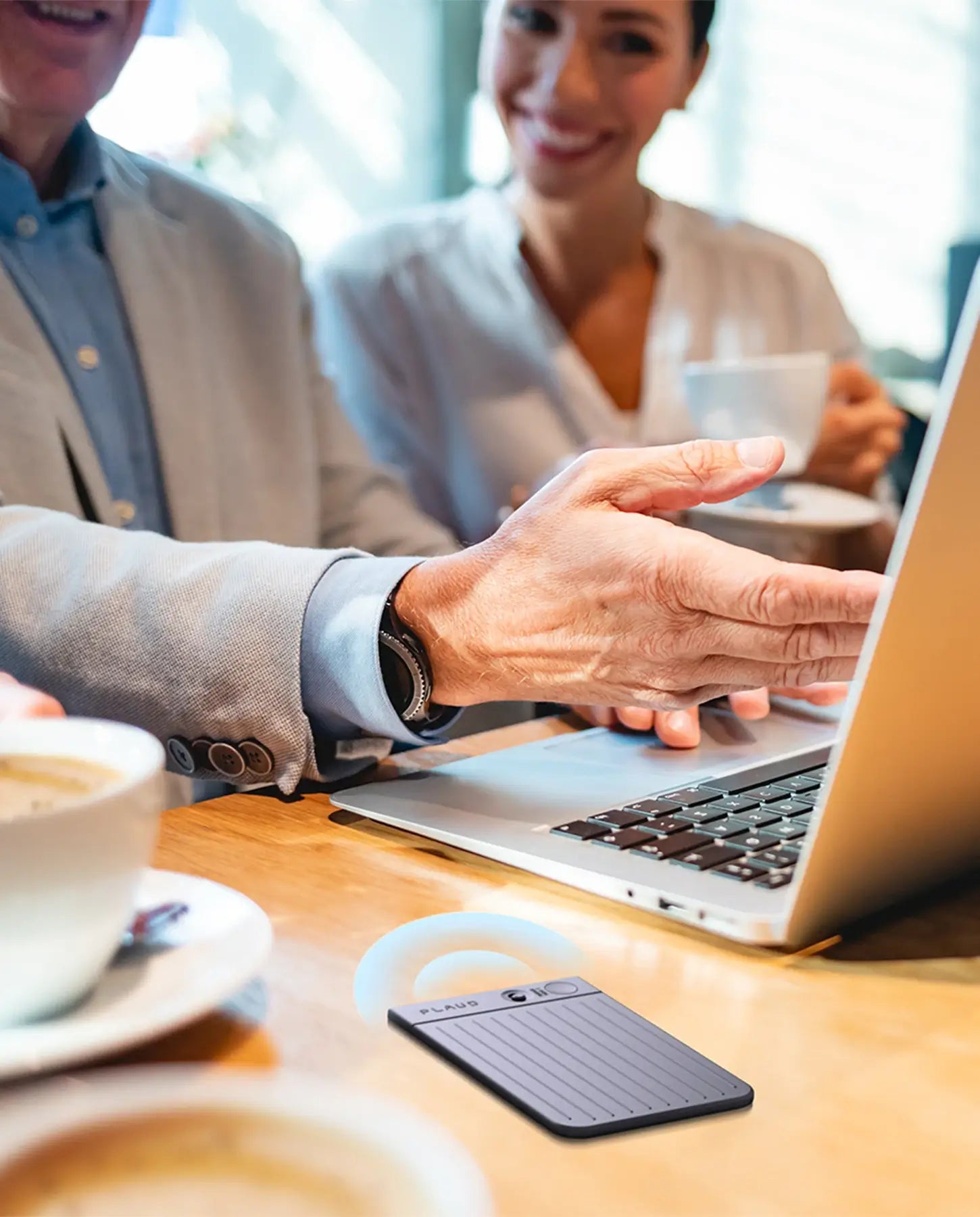 A grey Plaud Note records a discussion between two professionals at a cafe table, with one pointing at a laptop screen.