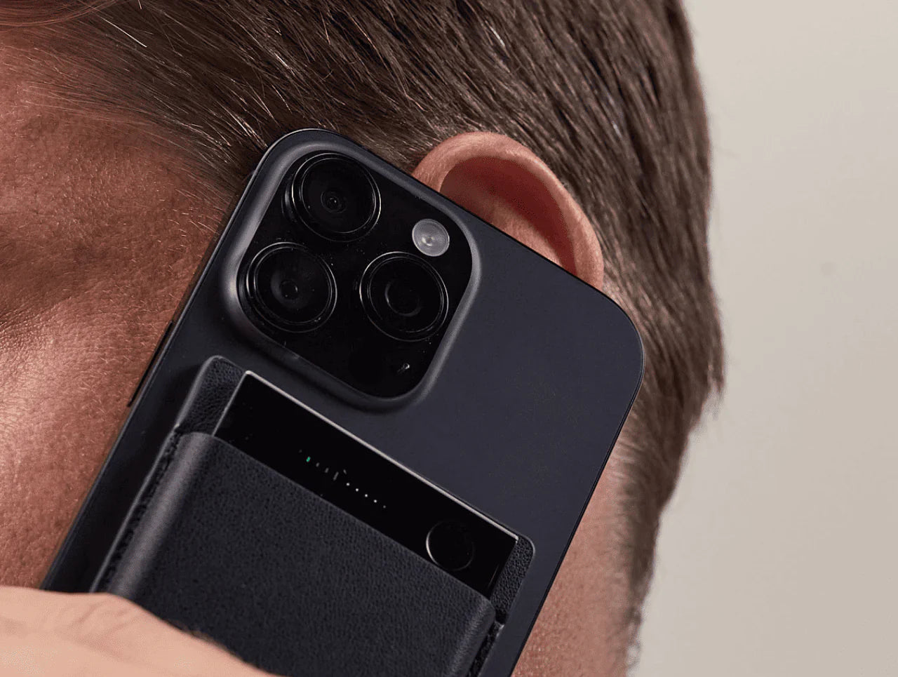 A black Plaud Note Pro, integrated into a black leather phone case, is held against a person's ear with a smartphone.