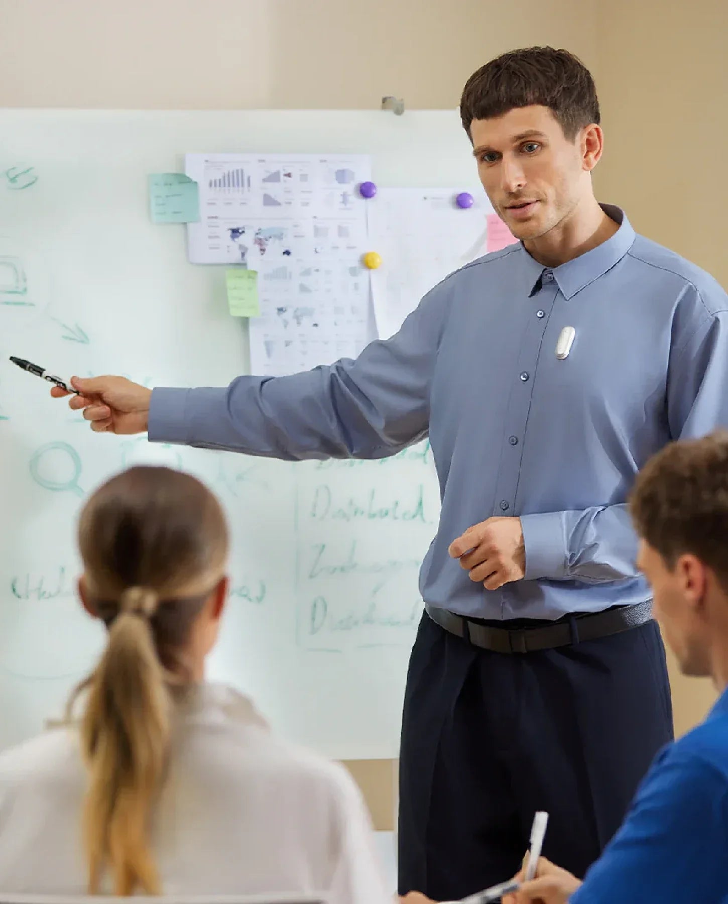 A man in a blue shirt presents to two people in front of a whiteboard, wearing a white Plaud NotePin S on his shirt.