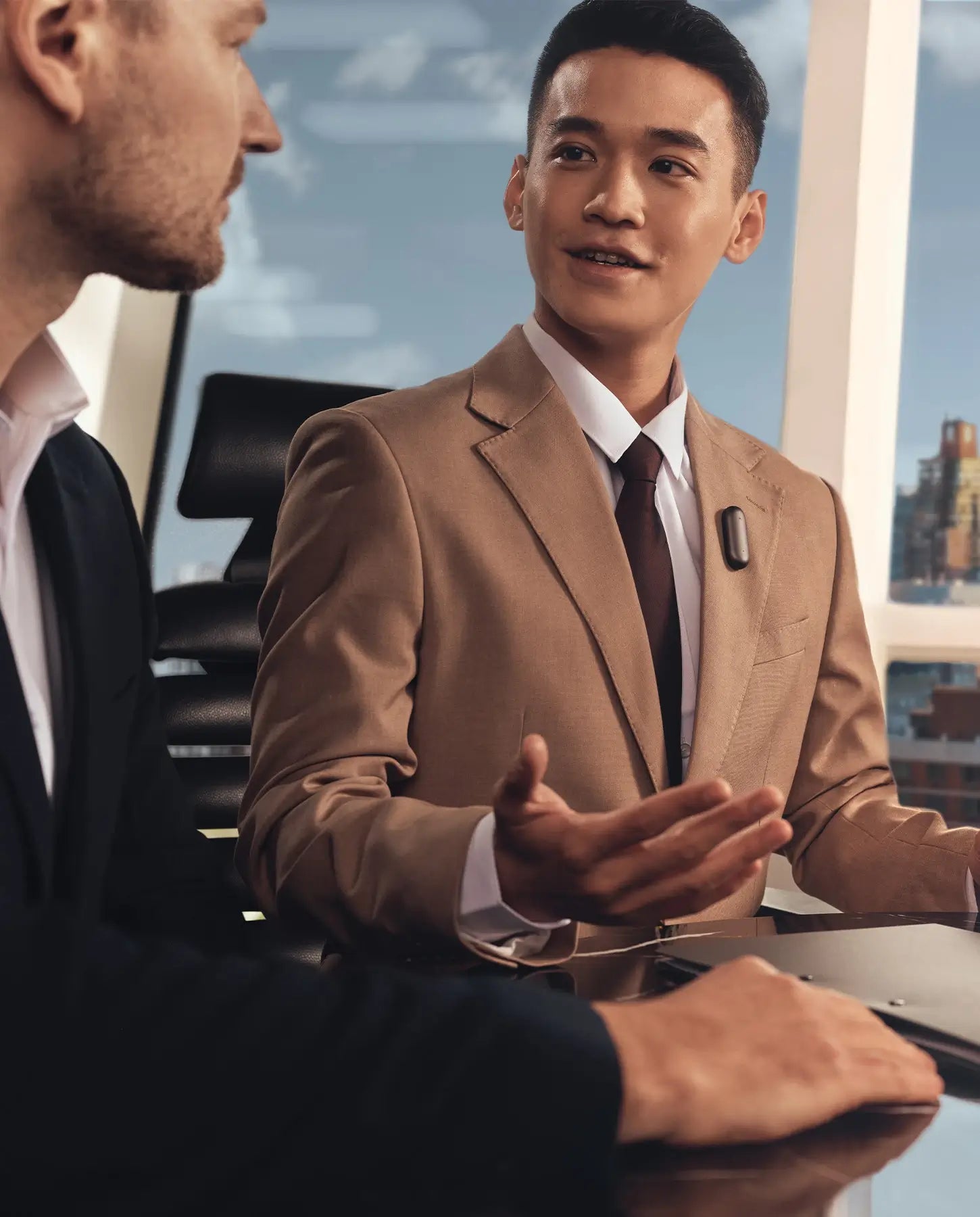 Two businessmen in suits discuss in an office with a city view, one wearing a black Plaud NotePin on his lapel.