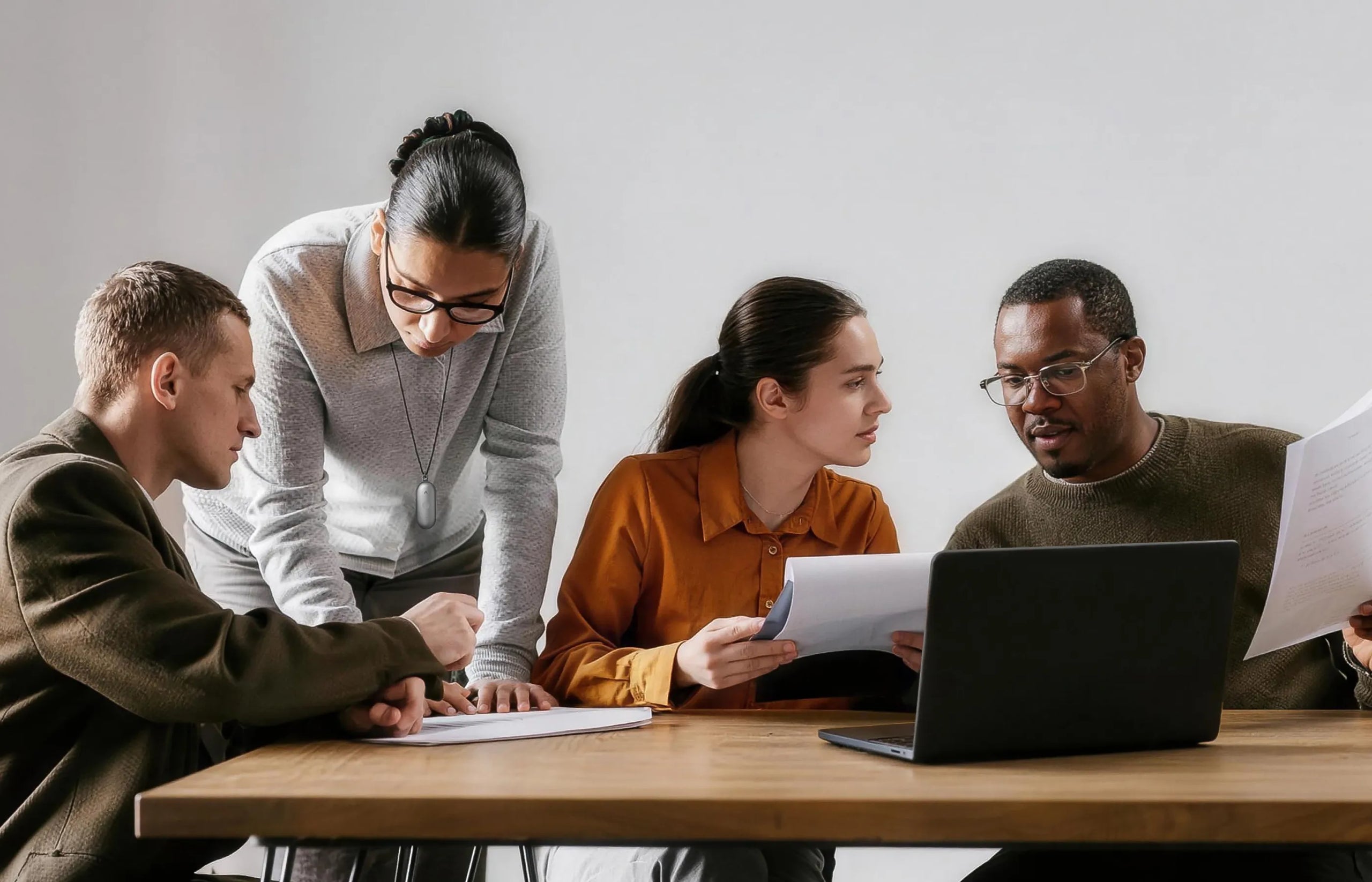 Four people discuss documents around a wooden table, with one woman wearing a white Plaud NotePin S on a lanyard.