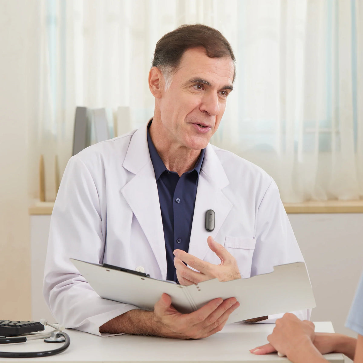 A male doctor in a white lab coat, wearing a black Plaud NotePin S on his chest, speaks to a patient while holding a fold