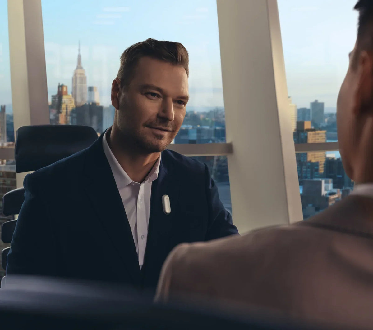 A man in a dark suit wears a white Plaud NotePin on his lapel while conversing with another person in a modern office overloo