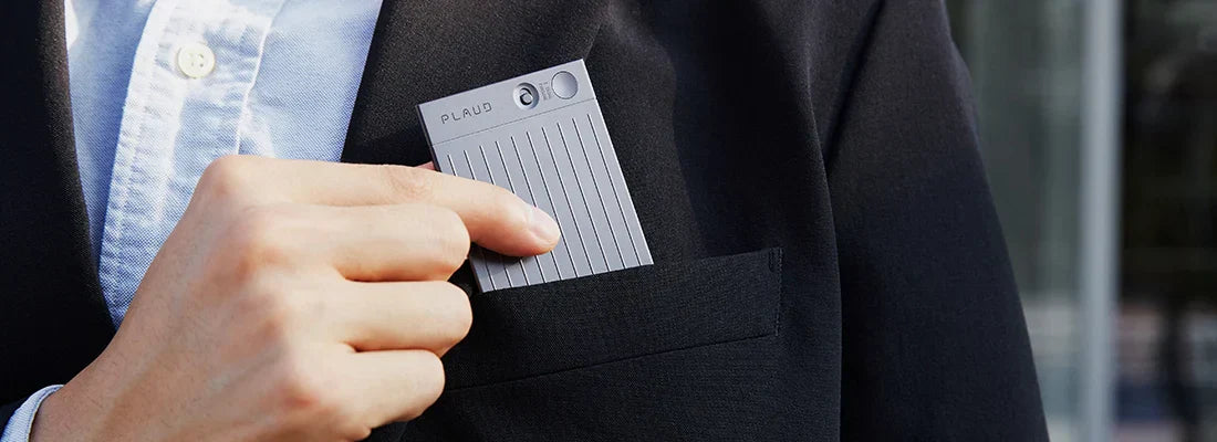 A hand slides a gray Plaud Note AI voice recorder into the breast pocket of a black suit jacket over a light blue shirt.