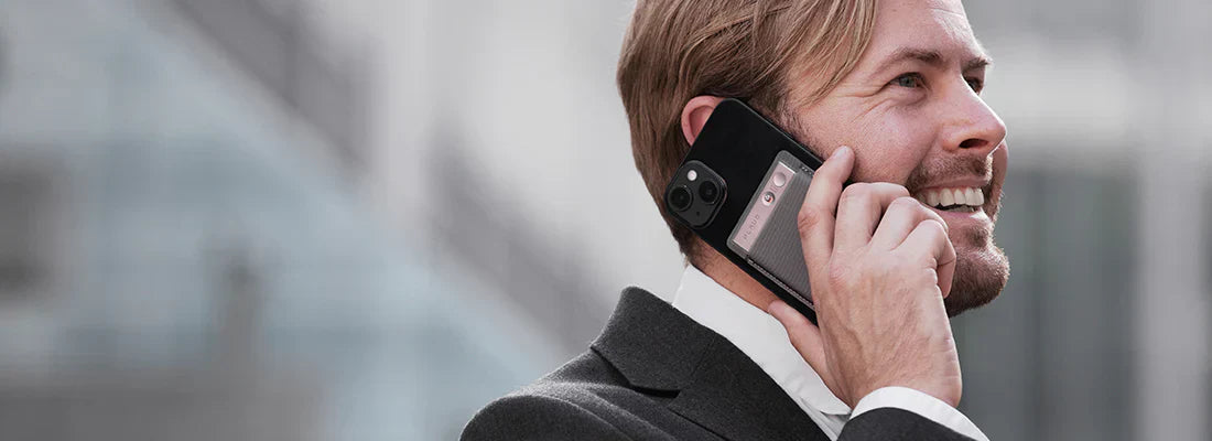 A smiling man in a suit talks on a black smartphone with a black Plaud Note Pro attached to its back.