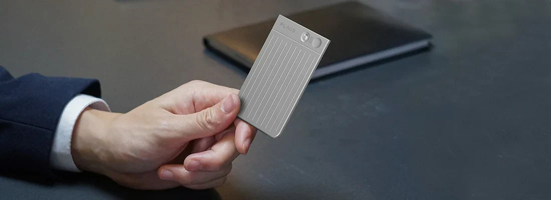 A hand in a dark suit holds a silver Plaud Note, showcasing its ribbed design and brushed-metal top, on a dark desk with a no