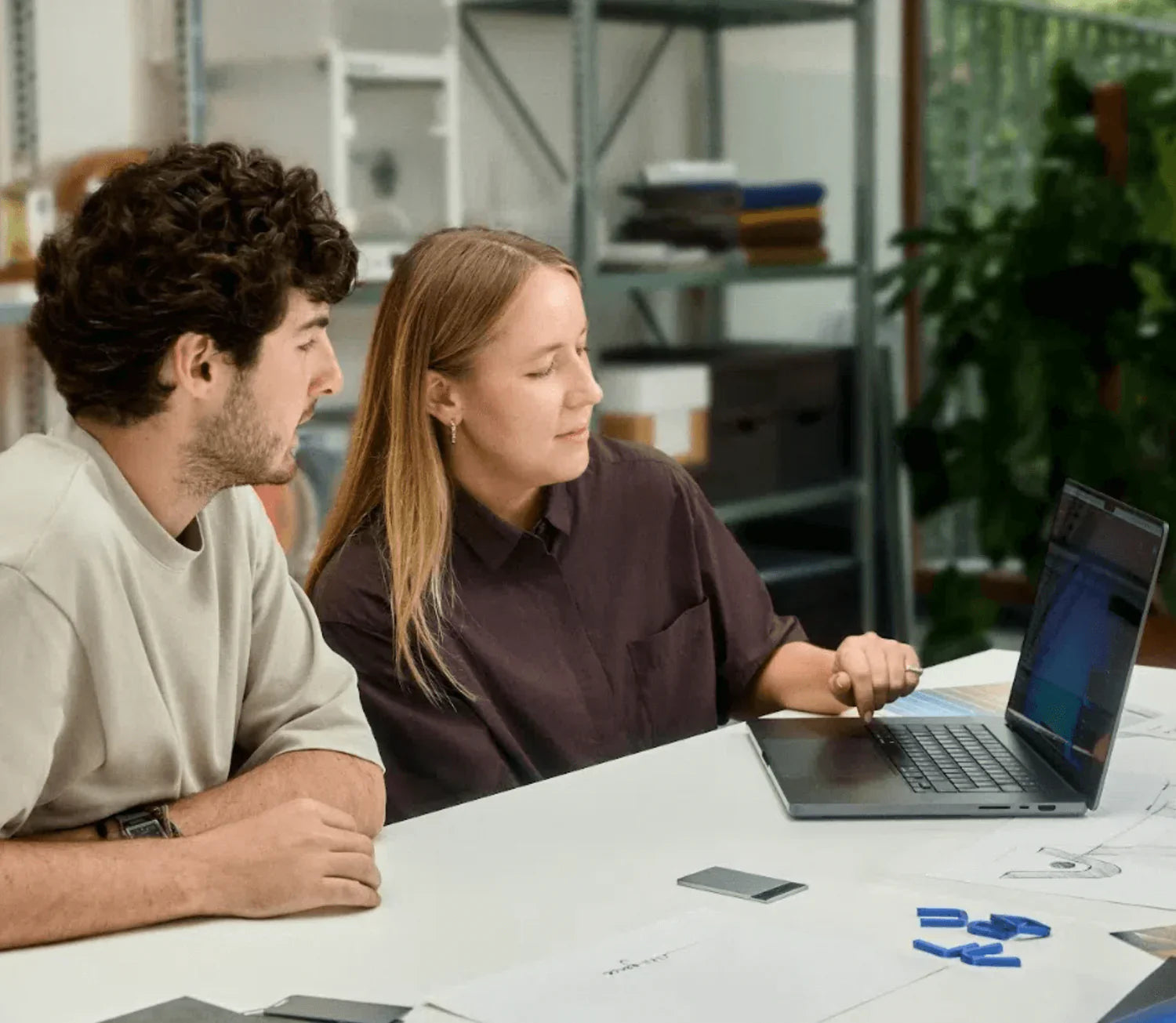 Two colleagues, a man and a woman, collaborate at a white table, looking at a laptop screen with a dark grey Plaud Note Pro n