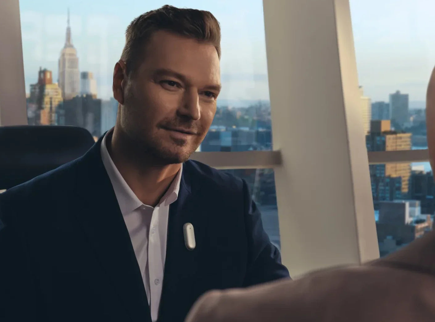 A man in a suit smiles during a meeting, wearing a white Plaud NotePin on his lapel, with a city skyline in the background.