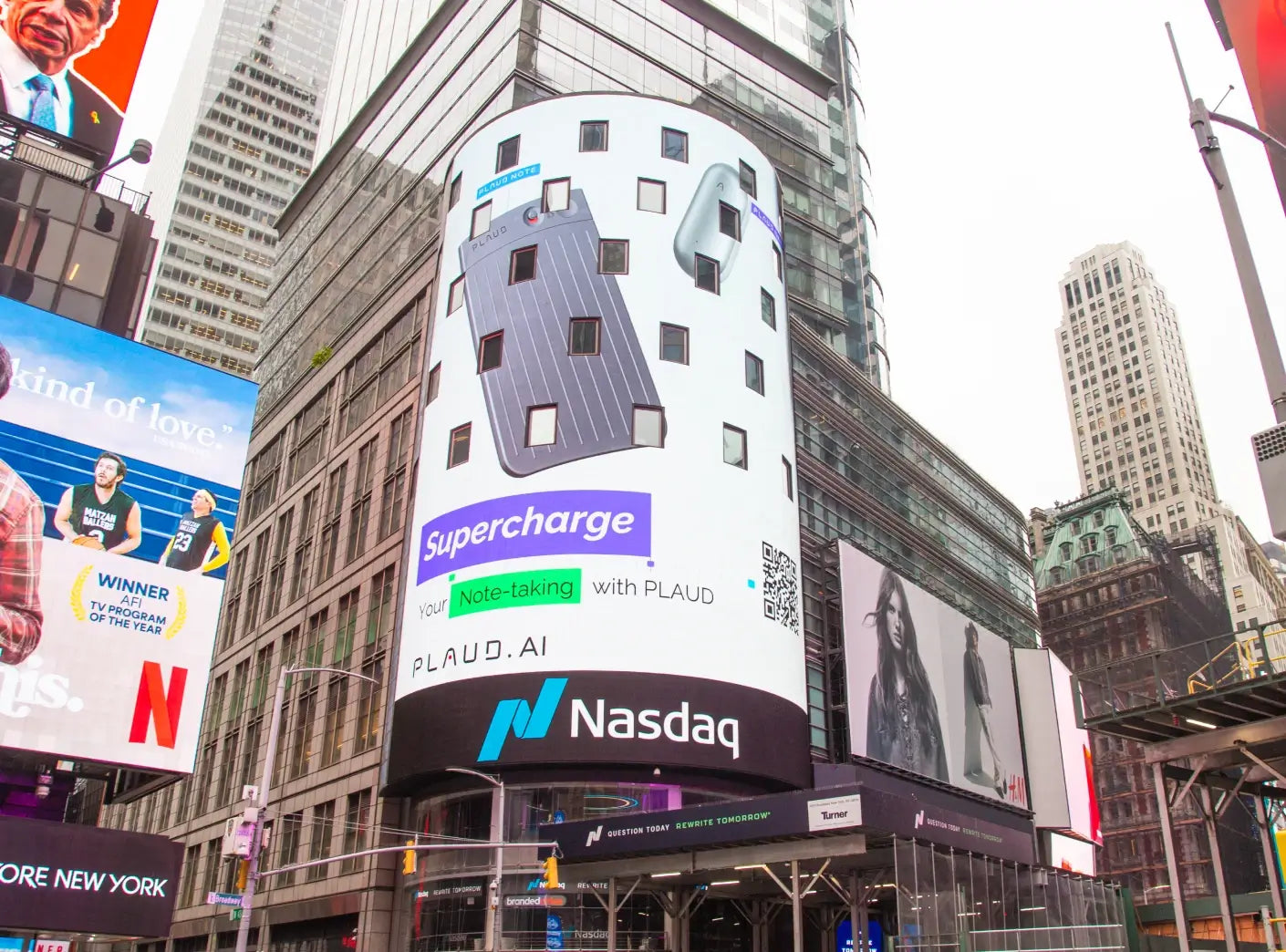 A large digital billboard in a bustling city displays a grey Plaud Note Pro and Plaud NotePin, advertising "Supercharge Your