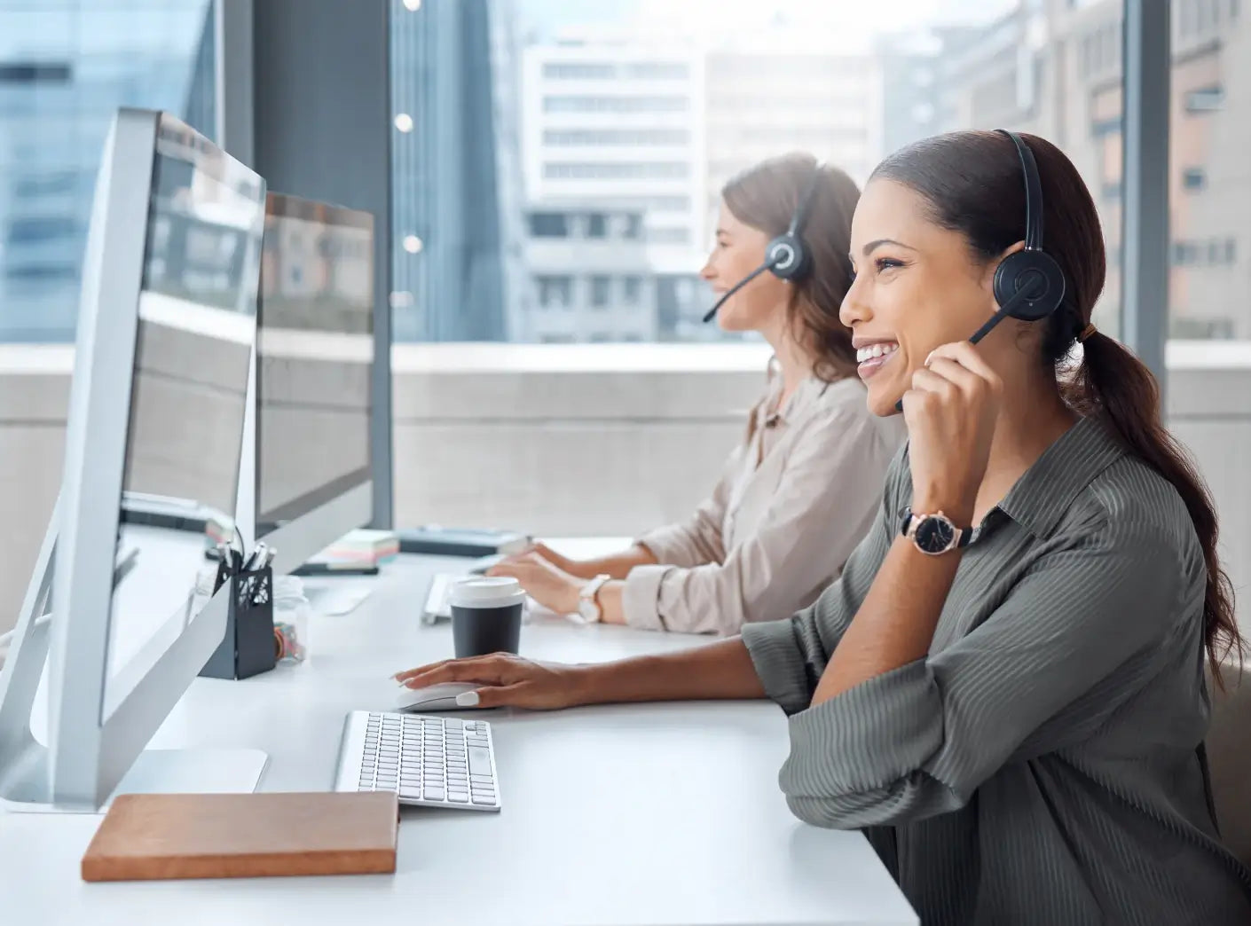 Two smiling women wearing headsets work at computers in a bright, modern office, conveying customer service.